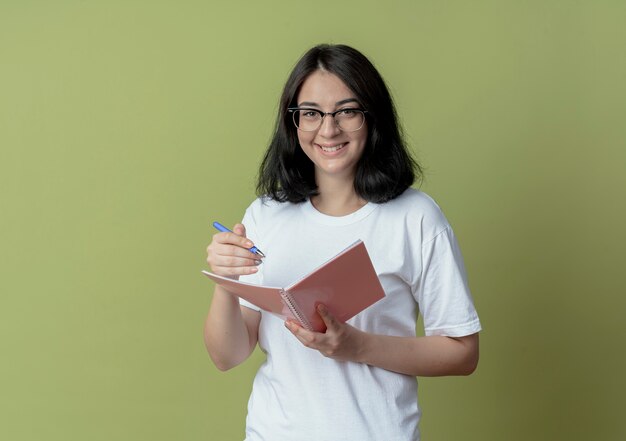 Mulher jovem e bonita caucasiana sorridente, usando óculos, segurando uma caneta e um bloco de notas isolado em um fundo verde oliva com espaço de cópia