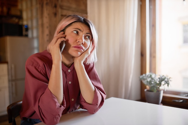 Mulher jovem e bonita com argola no nariz e cabelo rosa sentada à mesa, falando ao telefone