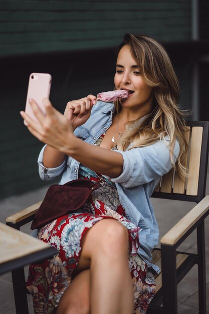 Mulher jovem e bonita com cabelo comprido come sorvete e faz selfie no smartphone. close-up retrato do lado de fora