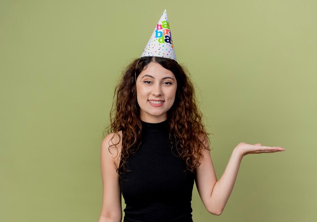 Mulher jovem e bonita com cabelo encaracolado com um boné de férias apresentando-se com um braço da mão sorrindo conceito de festa de aniversário em pé sobre a parede laranja