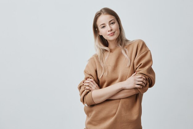 mulher jovem e bonita com cabelos loiros lisos, sorrindo gentilmente enquanto ouve uma conversa interessante, vestindo blusa de mangas compridas solta, mantendo os braços cruzados.