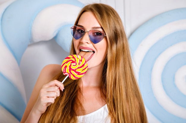 Mulher jovem e bonita com cabelos longos incríveis lambendo o pirulito grande, conceito de nutrição da dieta do açúcar doce, cores pastel dos lábios bonitos.