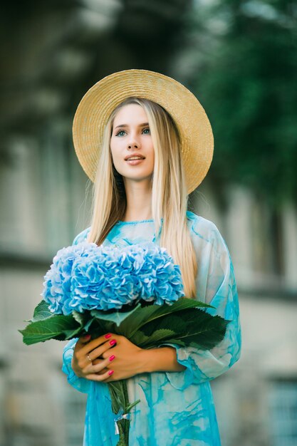 Mulher jovem e bonita com chapéu de palha sorrindo com hortênsia na rua do verão