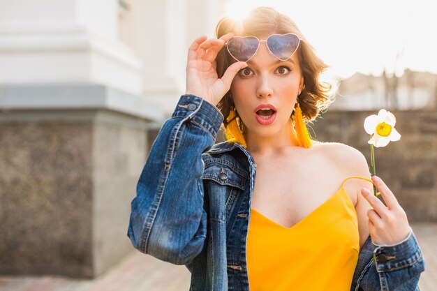 Mulher jovem e bonita com expressão de rosto surpreso, emocional, emoção chocada, vestindo roupas elegantes, jaqueta jeans, blusa amarela, segurando uma flor, verão ensolarado, óculos de sol engraçados da moda