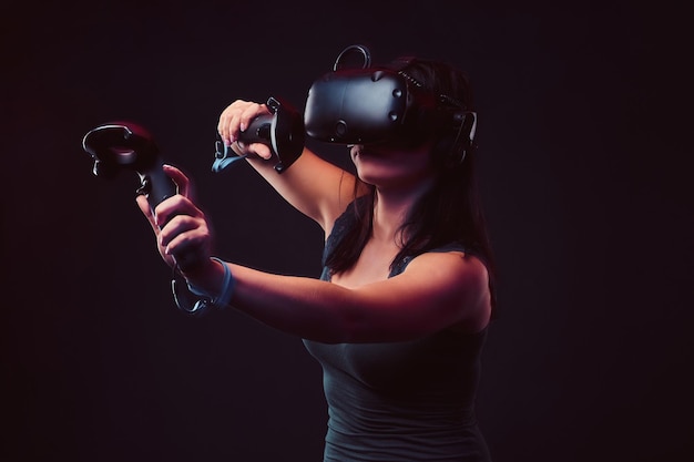 Mulher jovem e bonita com fone de ouvido de realidade virtual e joysticks jogando videogame. Isolado em fundo escuro.