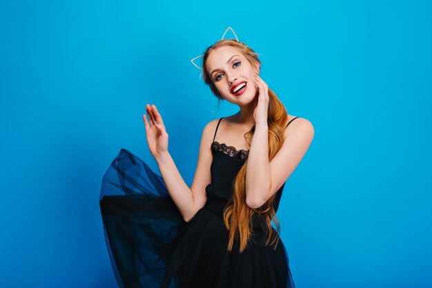 Mulher jovem e bonita com um lindo sorriso, vestido preto esvoaçante, posando. Ela tem cabelo comprido, usa bandana com orelhas de gato, bela maquiagem com batom vermelho.