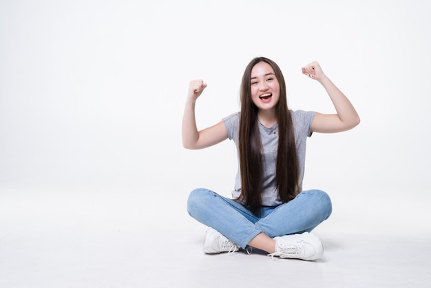 Mulher jovem e bonita comemorando o sucesso sentada no chão, isolada na parede branca