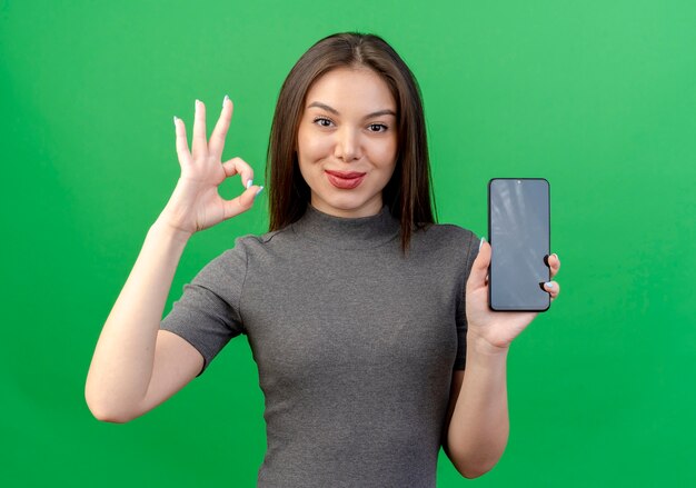 Mulher jovem e bonita confiante segurando um telefone celular e fazendo sinal de ok isolado no fundo verde