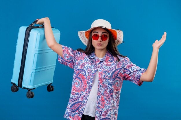 Mulher jovem e bonita confusa com chapéu de verão e óculos de sol vermelhos segurando uma mala de viagem.
