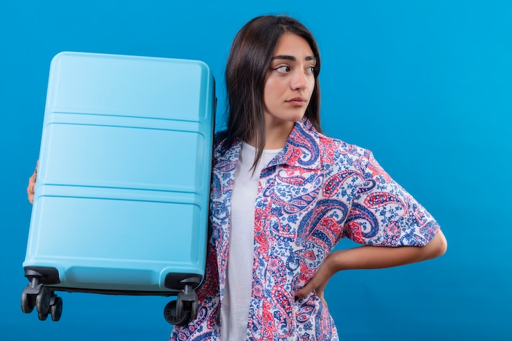 Foto grátis mulher jovem e bonita confusa, segurando uma mala de viagem, olhando para o lado com uma expressão facial duvidosa, parada sobre o espaço azul isolado