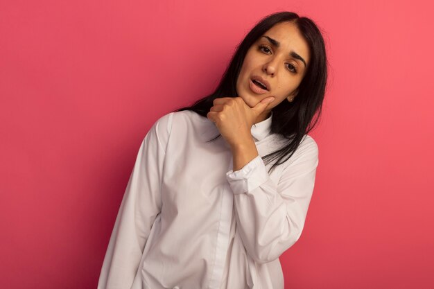 Mulher jovem e bonita descontente vestindo camiseta branca e colocando a mão no queixo