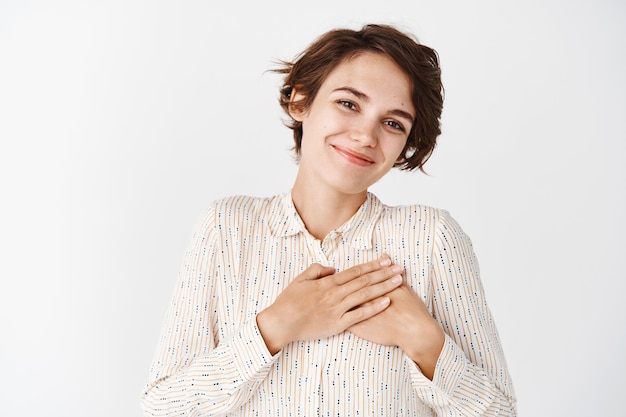 Mulher jovem e bonita dizendo obrigado, sorrindo e parecendo grata, segurando o coração com as mãos em agradecimento, em pé sobre uma parede branca