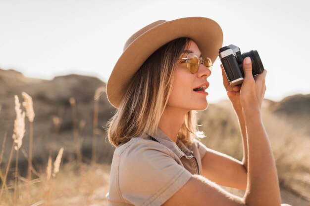 Mulher jovem e bonita e elegante em um vestido cáqui no deserto, viajando pela África em um safári, usando um chapéu, tirando foto com uma câmera vintage