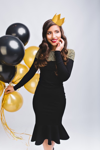 Mulher jovem e bonita elegante na moda vestido para celebrar a festa de ano novo, segurando balões dourados e pretos. Tem cabelo castanho comprido, coroa amarela. Se divertindo, noite mágica, aniversário.