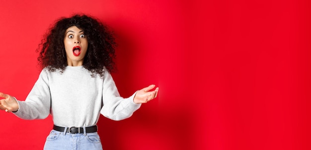 Foto grátis mulher jovem e bonita em pé com o queixo caído e olhando impressionado para a câmera espalhar as mãos para o lado