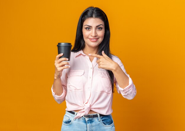 Mulher jovem e bonita em roupas casuais segurando uma xícara de café apontando com o dedo indicador para ela e sorrindo confiante em pé sobre a parede laranja