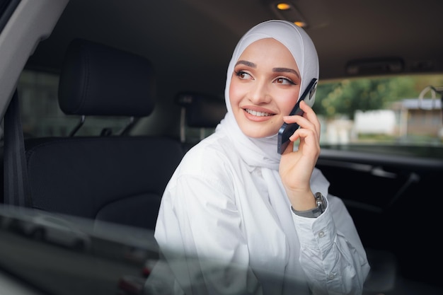 Mulher jovem e bonita em um hijab sentada em um carro e falando ao telefone