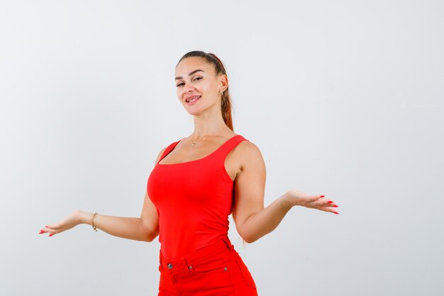 Mulher jovem e bonita em um top vermelho, calças mostrando um gesto de boas-vindas e olhando alegre, vista frontal.