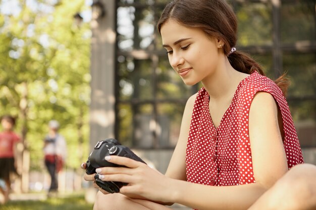 Foto grátis mulher jovem e bonita em um vestido de polca vermelha sentada ao ar livre com uma câmera dslr, aprendendo a tirar fotos profissionais, rolando fotos, olhando para a tela