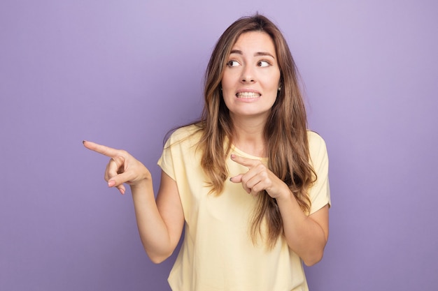 Mulher jovem e bonita em uma camiseta bege olhando para o lado confusa, apontando com os dedos indicadores para o lado em pé sobre o roxo