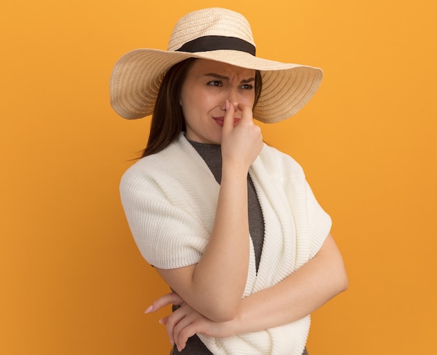 Mulher jovem e bonita enojada com chapéu de praia fazendo gesto de mau cheiro olhando para o lado isolado na parede laranja