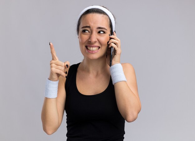 Mulher jovem e bonita esportiva sorridente, usando bandana e pulseiras, falando no telefone, olhando para o lado apontando para cima, isolado na parede branca com espaço de cópia