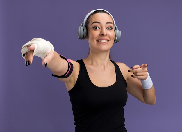 Mulher jovem e bonita esportiva sorridente usando bandana, pulseiras, fones de ouvido e uma braçadeira de telefone com o pulso ferido envolto em bandagem, olhando e apontando para a frente, isolado na parede roxa