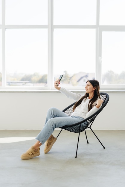 Mulher jovem e bonita fazendo selfie pelo telefone e sorrindo enquanto está sentado na cadeira grande em casa
