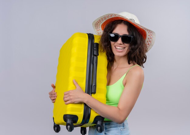 Mulher jovem e bonita feliz com chapéu e óculos escuros, segurando a mala na parede branca isolada