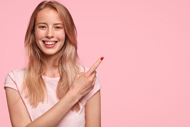 Mulher jovem e bonita feliz com expressão positiva alegre, vestida com uma camiseta casual rosa claro, indica no canto superior direito