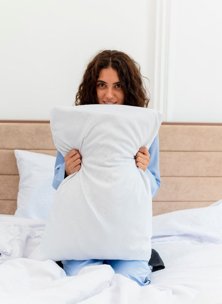 Foto grátis mulher jovem e bonita feliz de pijama azul, sentada na cama com travesseiro e sorriso no rosto, no interior do quarto
