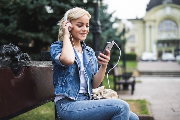 Mulher jovem e bonita feliz ouvindo música em fones de ouvido e usando o smartphone enquanto está sentado no banco da cidade