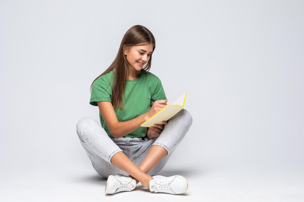 Mulher jovem e bonita feliz vestindo roupas casuais, sentada no chão com as pernas cruzadas isoladas em branco, segurando um diário