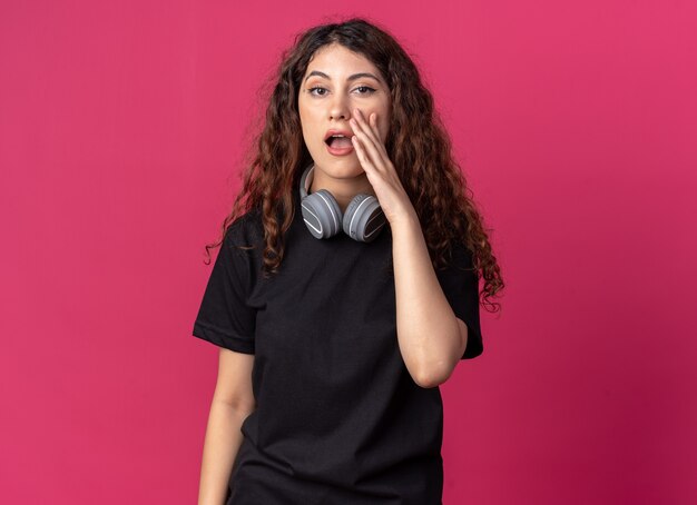 Mulher jovem e bonita impressionada usando fones de ouvido no pescoço e tocando o rosto