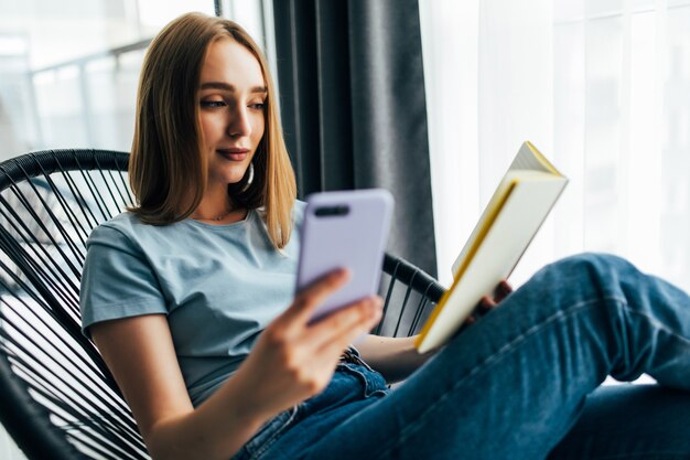 Mulher jovem e bonita lendo um livro e sentada em uma cadeira confortável em casa