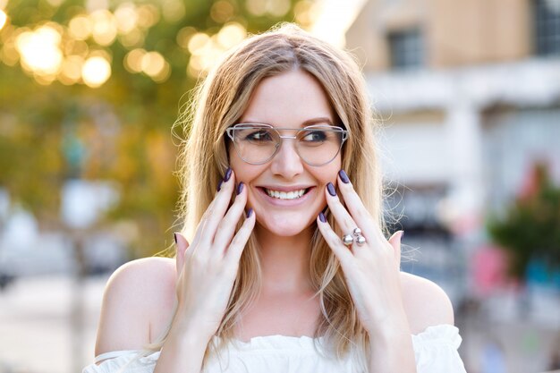 Mulher jovem e bonita loira sorrindo e fazendo careta, óculos transparentes e camisa branca, posando no parque da cidade