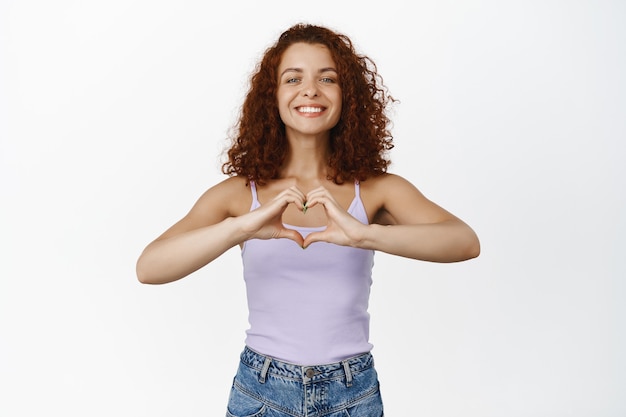 Foto grátis mulher jovem e bonita mostrando o sinal do coração e sorrindo, vestindo a blusa de verão em branco.