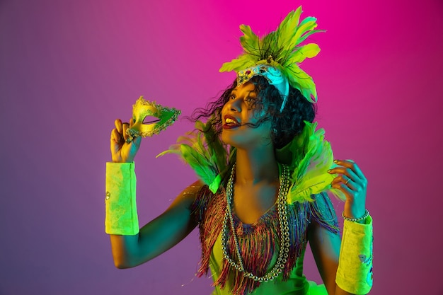 Mulher jovem e bonita no carnaval, elegante fantasia de baile de máscaras com penas dançando na parede gradiente em neon