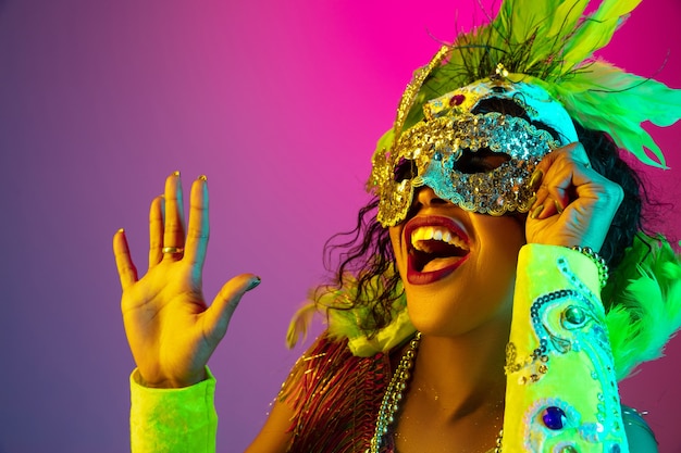 Mulher jovem e bonita no carnaval, fantasia elegante de baile de máscaras com penas na parede gradiente em luz de néon