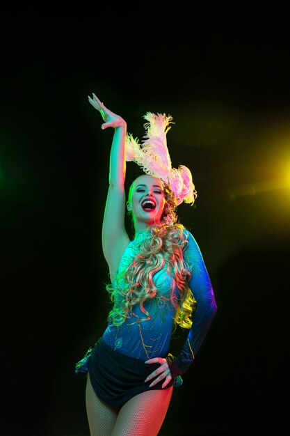 Mulher jovem e bonita no carnaval, fantasia elegante de baile de máscaras com penas na parede preta com luz neon