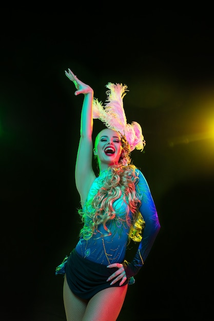 Mulher jovem e bonita no carnaval, fantasia elegante de baile de máscaras com penas na parede preta com luz neon