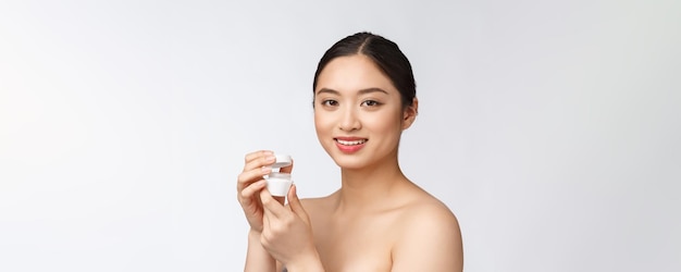 Mulher jovem e bonita no fundo branco isolado segurando o creme facial cosmético asiático