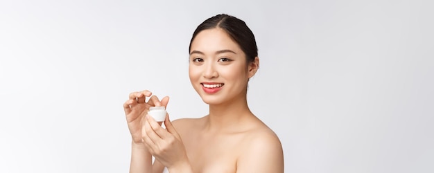 Mulher jovem e bonita no fundo branco isolado segurando o creme facial cosmético asiático