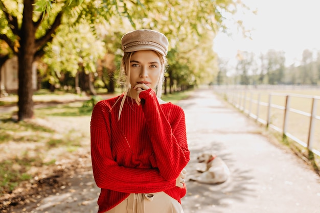 Mulher jovem e bonita no suéter vermelho e bonito chapéu moderno parecendo pensativo no outono park. loira atraente em roupas elegantes posando ao ar livre.