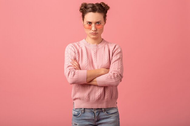 Mulher jovem e bonita pensando tendo um problema, olhando para baixo com um suéter rosa e óculos de sol, isolados no fundo rosa do estúdio