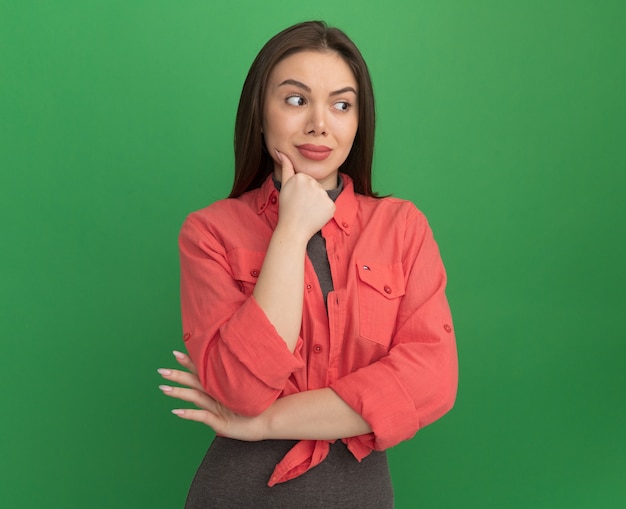 Mulher jovem e bonita pensativa colocando a mão no queixo, olhando para o lado isolado na parede verde com espaço de cópia