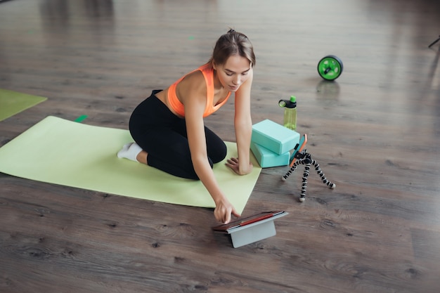 Mulher jovem e bonita praticando ioga, está envolvida com o professor online através de um tablet. Conceito de esportes em casa.