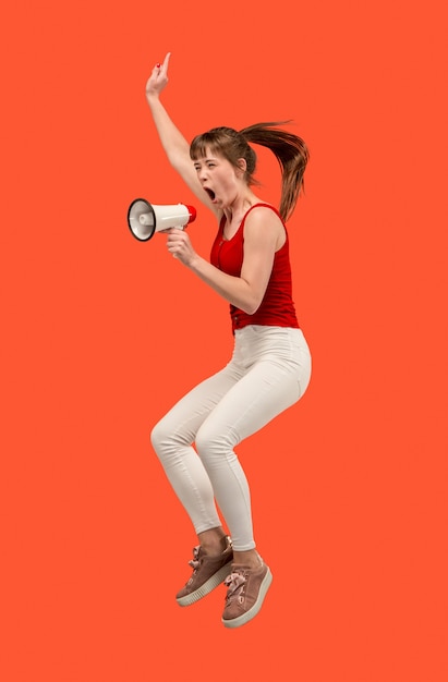 Mulher jovem e bonita pulando com o megafone isolado sobre fundo vermelho. Garota correndo em movimento ou movimento. Emoções humanas e conceito de expressões faciais