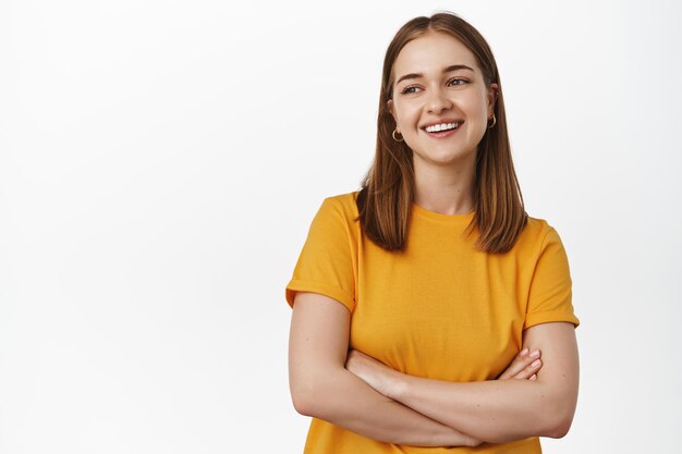 Mulher jovem e bonita segura os braços cruzados no peito, rindo e sorrindo, olhando de lado o texto promocional no espaço vazio esquerdo, publicidade, de pé na camiseta amarela contra fundo branco.