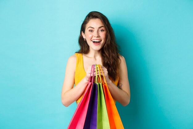 Mulher jovem e bonita segurando sacolas de compras, sorrindo animada para a câmera, comprando com descontos, em pé sobre um fundo azul